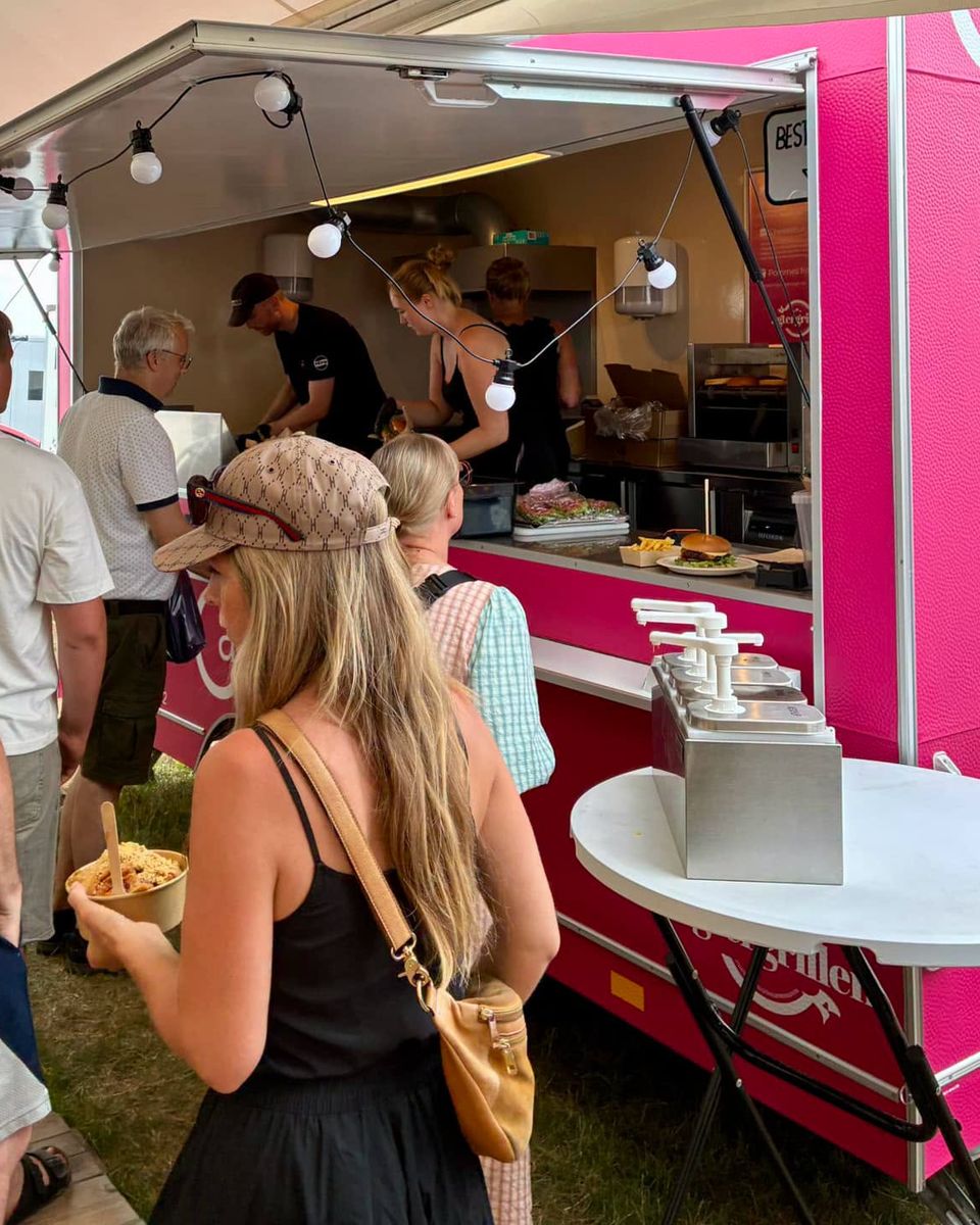 Gæster i kø ved den pink Carlsvognen under et festivaltelt med lyskæder